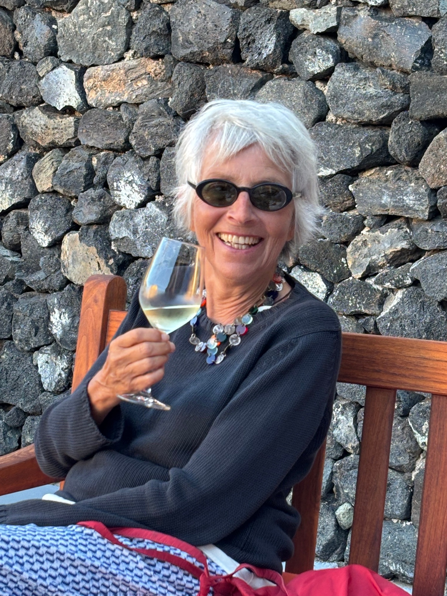 Smiling woman with white hair enjoying a glass of white wine in front of a lava stone wall