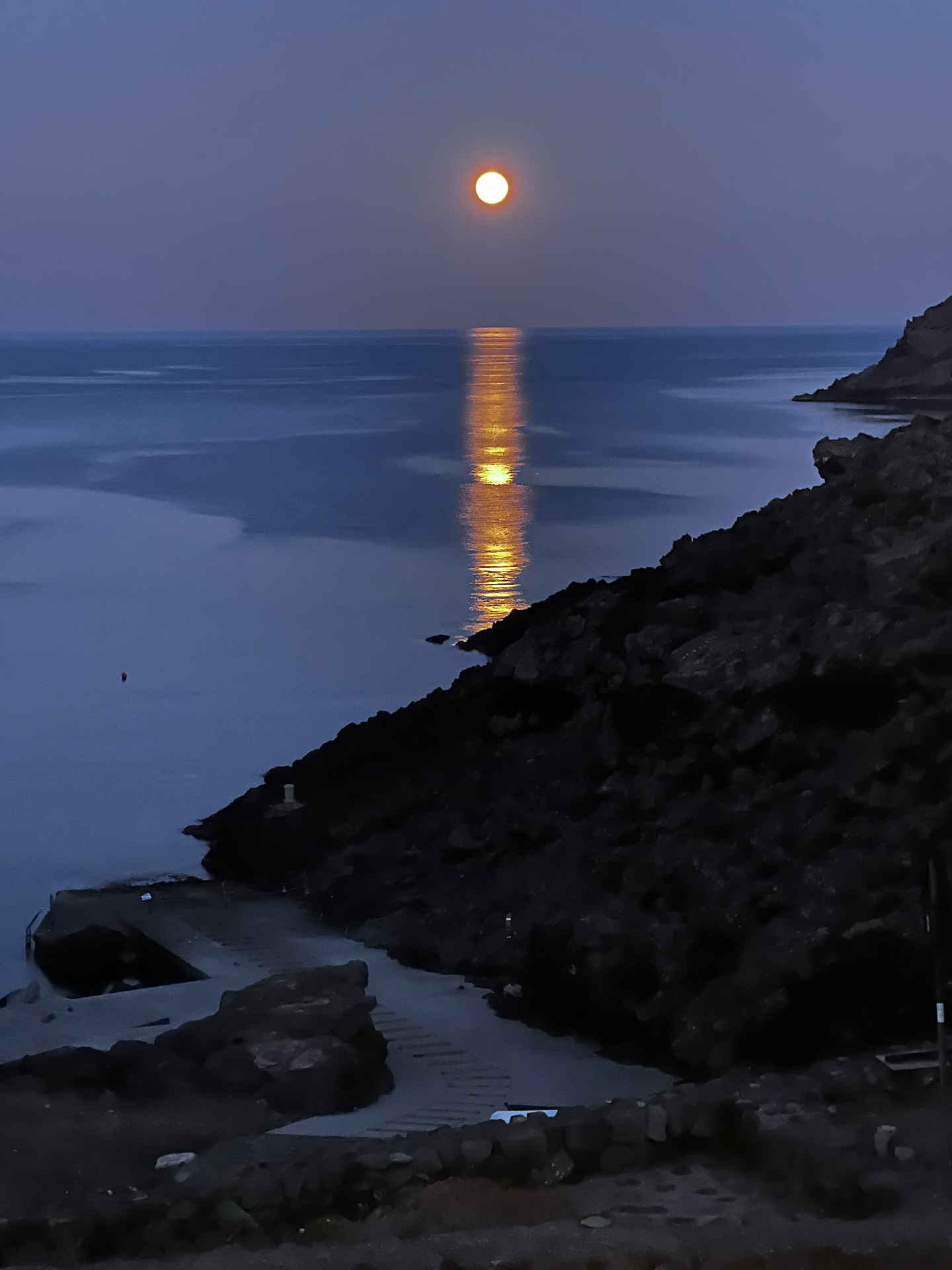 Full moon reflecting across a calm sea beside dark volcanic cliffs at dusk